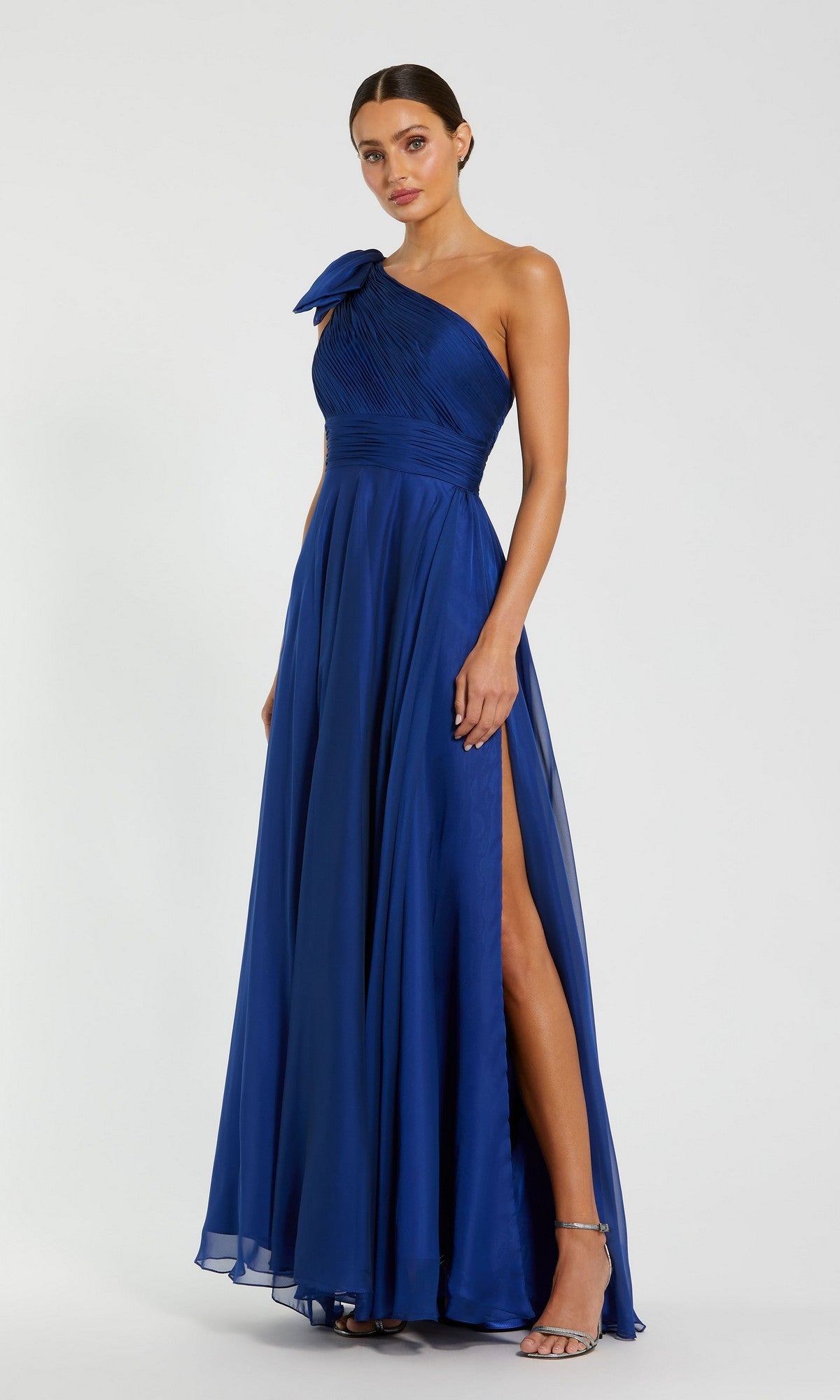 A woman models the Mac Duggal 67476 long one-shoulder royal blue prom dress with a bow detail and thigh-high slit, standing against a plain background.